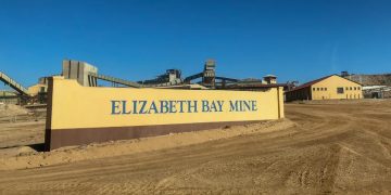 Entrance sign for Elizabeth Bay Mine with yellow wall and blue lettering, industrial buildings in the background under a clear blue sky.