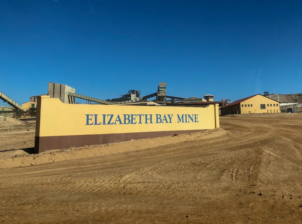 Entrance sign for Elizabeth Bay Mine with yellow wall and blue lettering, industrial buildings in the background under a clear blue sky.