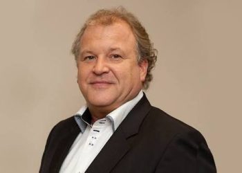 Duane Parnham1 | Mining and Energy Namibia | Namibia’s Leading Mining & Energy News Portrait of a middle-aged man in a dark suit and white shirt, smiling against a beige background.