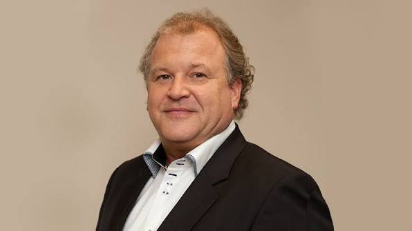 Portrait of a middle-aged man in a dark suit and white shirt, smiling against a beige background.