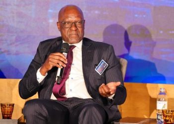Man in a dark pinstripe suit and maroon tie speaks into a handheld microphone on stage at a conference, wearing a name badge.