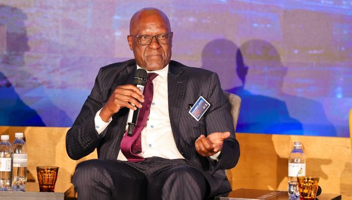 Man in a dark pinstripe suit and maroon tie speaks into a handheld microphone on stage at a conference, wearing a name badge.