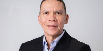 Professional headshot of a middle-aged man in a dark blazer and light blue shirt against a light gray background, smiling subtly.
