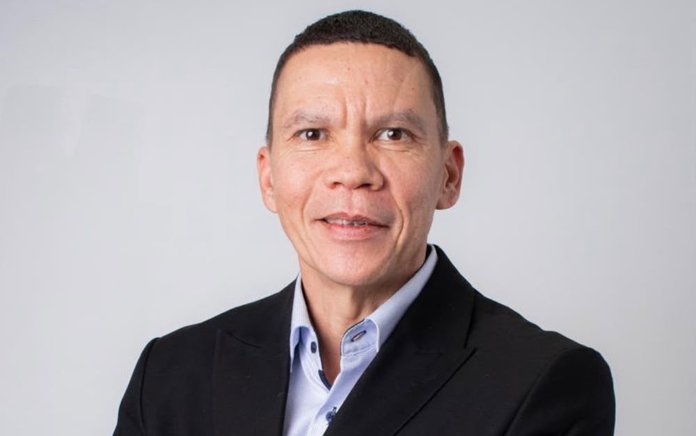 Professional headshot of a middle-aged man in a dark blazer and light blue shirt against a light gray background, smiling subtly.