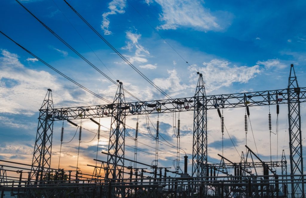 Electrical substation with metal towers and power lines silhouetted against a sunset sky, cables crisscrossing overhead.