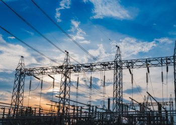 Electrical substation with metal towers and power lines silhouetted against a sunset sky, cables crisscrossing overhead.