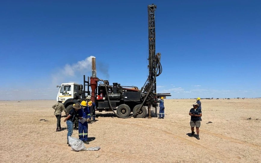 Mobile drilling rig mounted on a truck in a flat desert, workers in hard hats and coveralls nearby.