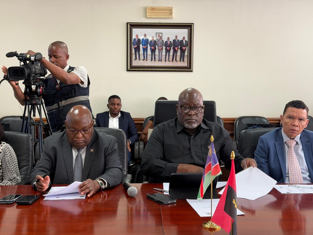 Camera operator films a formal meeting as officials sit at a polished table with flags and documents nearby.