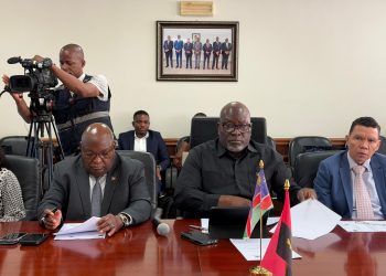 Camera operator films a formal meeting as officials sit at a polished table with flags and documents nearby.