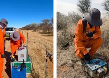 Aldoro-Kalkfeld | Mining and Energy Namibia | Namibia’s Leading Mining & Energy News Two researchers in bright orange coveralls and wide-brim hats set up field equipment in a dry savanna; one adjusts gear from a green/blue case, a vehicle in the background.