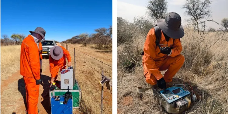 Two researchers in bright orange coveralls and wide-brim hats set up field equipment in a dry savanna; one adjusts gear from a green/blue case, a vehicle in the background.