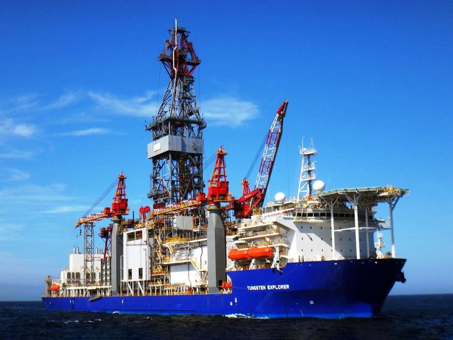 Offshore drilling ship Tungsten Explorer with tall derrick and red cranes at sea under a blue sky