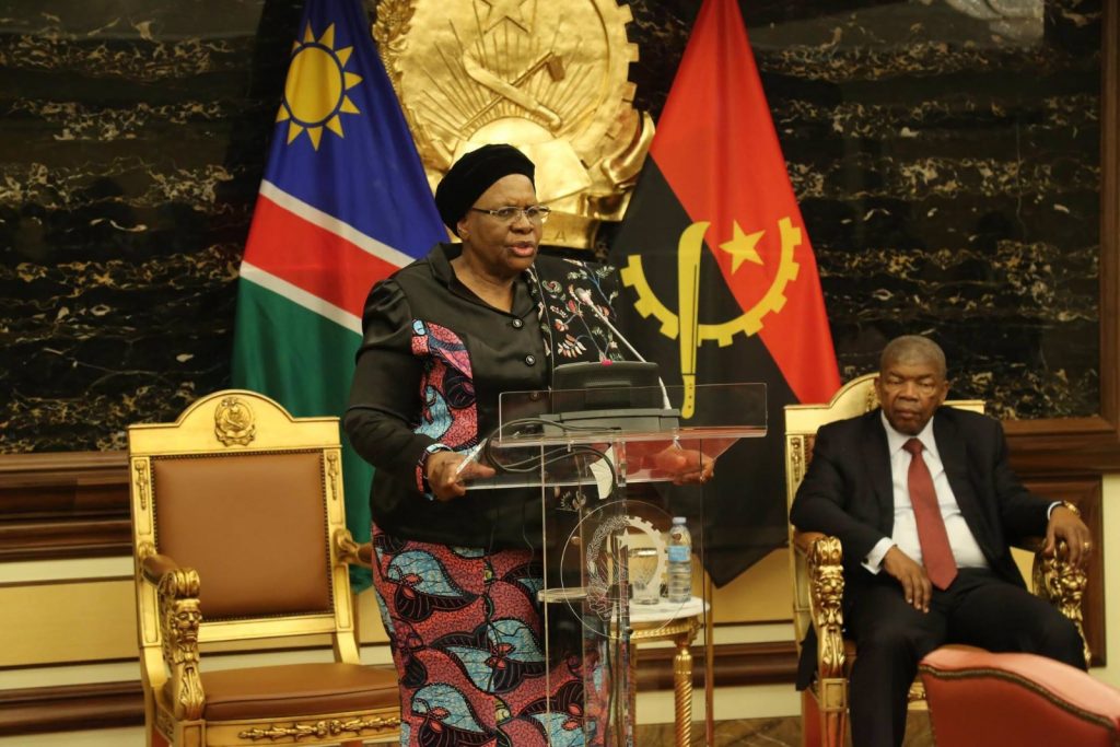 Woman in a patterned suit speaks at a clear podium during a formal government event, with flags behind her and a man seated nearby.