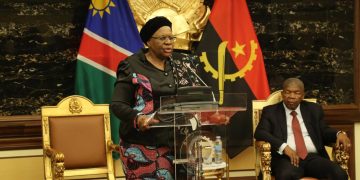 Woman in colorful patterned outfit giving a formal speech at a podium with two flags behind her and a man seated nearby on a golden chair in a formal hall