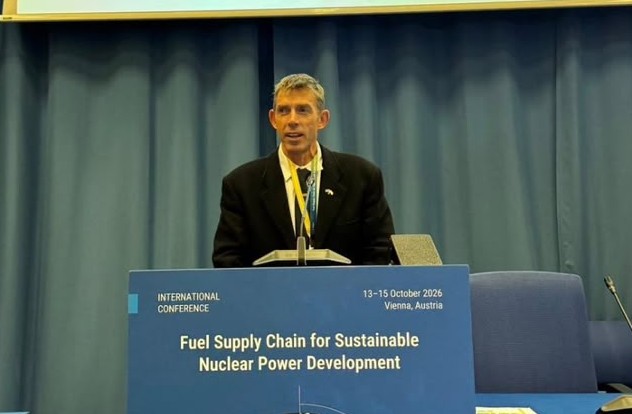 Male speaker in a black suit at a podium during an international conference; banner reads 'Fuel Supply Chain for Sustainable Nuclear Power Development'