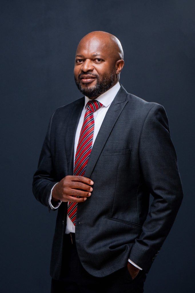 Professional portrait of a bald man with a beard in a dark gray suit and red striped tie, standing against a dark backdrop.