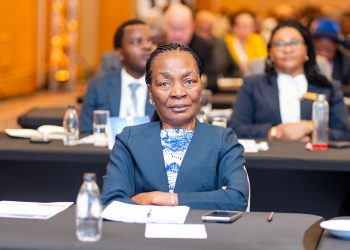 Front-on portrait of a professional woman in a blue blazer sitting at a conference table, with a blurred audience in the background.