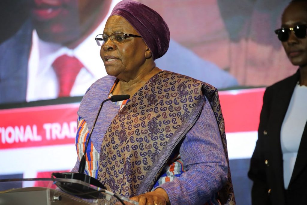 PRESIDENT4 | Mining and Energy Namibia | Namibia’s Leading Mining & Energy News Older woman in a purple headscarf and patterned dress speaks at a podium with a microphone at a conference, with a person in sunglasses in the background.