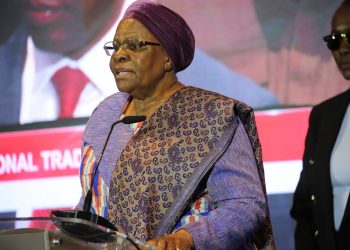 Older woman in a purple headscarf and patterned dress speaks at a podium with a microphone at a conference, with a person in sunglasses in the background.