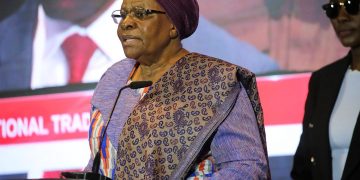 PRESIDENT4 | Mining and Energy Namibia | Namibia’s Leading Mining & Energy News Older woman in a purple headscarf and patterned dress speaks at a podium with a microphone at a conference, with a person in sunglasses in the background.