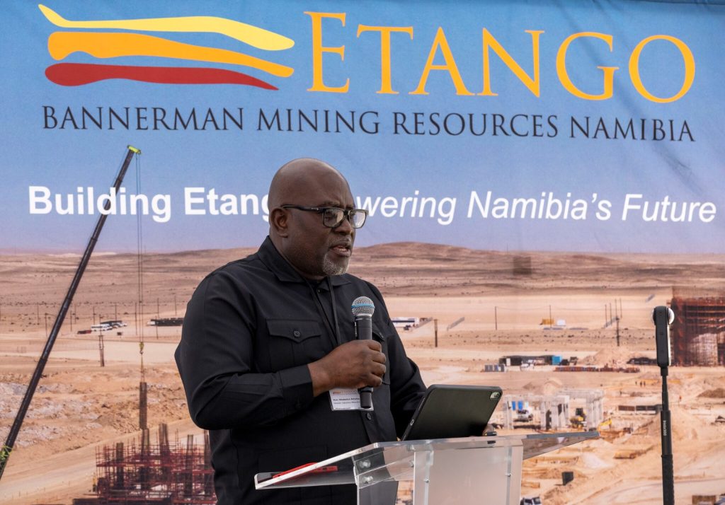 MODESTUS AMUTSE3 | Mining and Energy Namibia | Namibia’s Leading Mining & Energy News Male speaker wearing glasses and a black shirt delivers a talk at a podium in front of a large blue banner that reads ETANGO and Bannerman Mining Resources Namibia, set against a desert mining site backdrop.