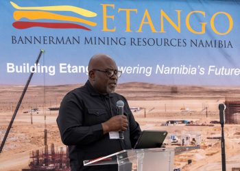 Male speaker wearing glasses and a black shirt delivers a talk at a podium in front of a large blue banner that reads ETANGO and Bannerman Mining Resources Namibia, set against a desert mining site backdrop.