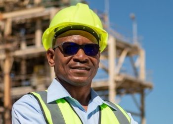GEORGE BOTSHIWE1 | Mining and Energy Namibia | Namibia’s Leading Mining & Energy News Construction worker wearing a neon yellow hard hat and safety vest at a building site with scaffolding in the background.