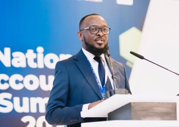 Man in a navy suit and glasses speaks at a podium during a conference, blue backdrop reading 'National Accounts Summit' behind him.
