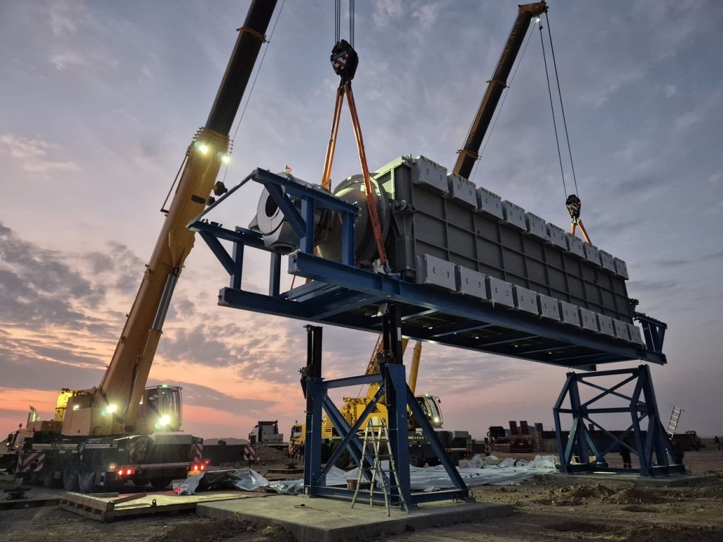 Two large cranes lift stacked concrete blocks onto a blue steel platform at a construction site during sunset.