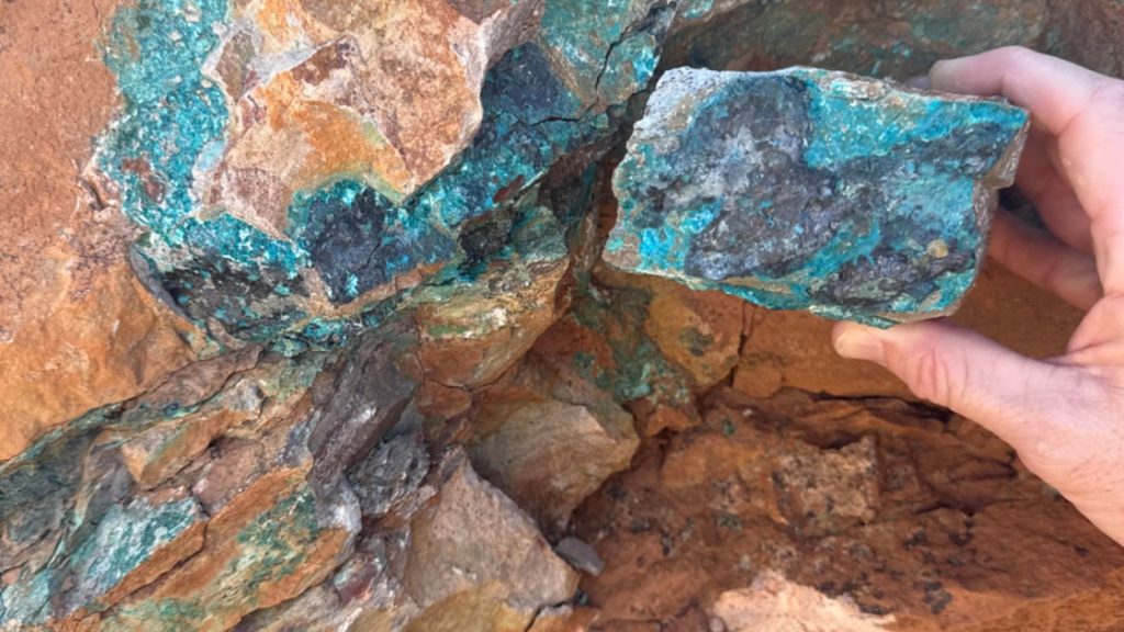 copper 5 | Mining and Energy Namibia | Namibia’s Leading Mining & Energy News Hand holding a rough rock with blue‑green copper minerals against a rusty rocky background underground/outcrop.