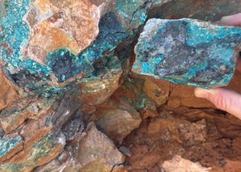 Hand holding a rough rock with blue‑green copper minerals against a rusty rocky background underground/outcrop.